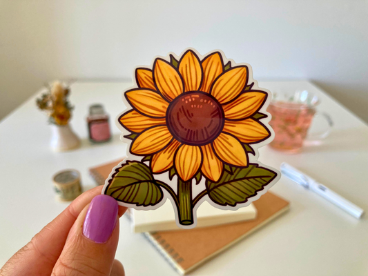 A hand with purple nail polish holds a cartoon sunflower sticker with bright yellow petals and green leaves. The background shows a desk with a notebook, pen, dried flowers, washi tape, a jar, and a glass cup of tea.