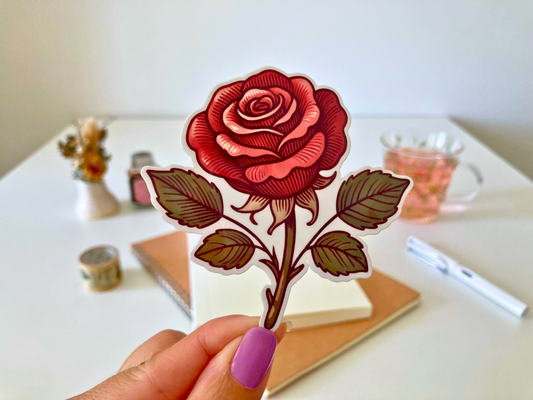 A hand with purple nail polish holds a cartoon sticker of a red rose with green leaves. The background features a desk with a notebook, pen, dried flowers, washi tape, a jar, and a glass cup of tea.