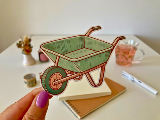 A hand with purple nail polish holds an anime-style sticker of a green wheelbarrow with brown outlines. The background features a white table with notebooks, a pen, a glass of pink tea, and decorative items.