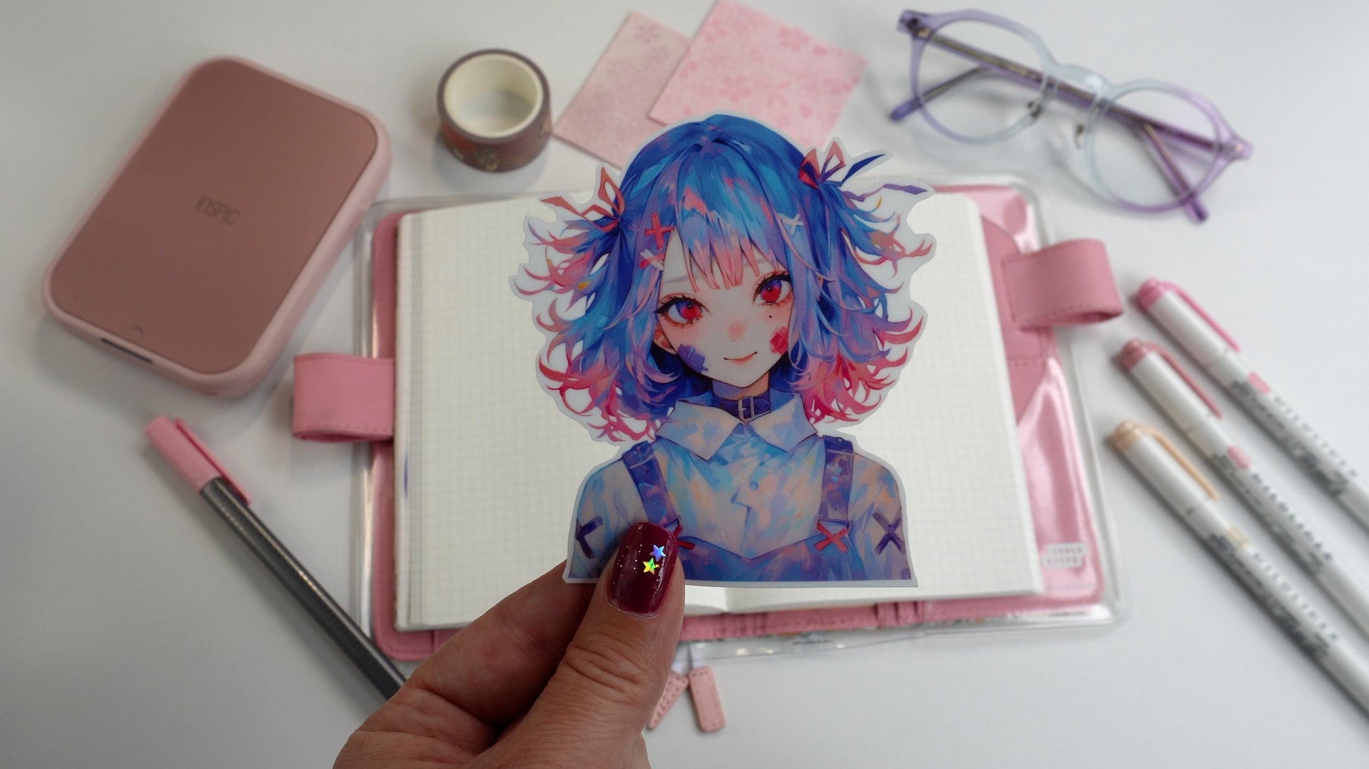 a hand holds a sticker of an anime girl with blue and red hair over a desk containing a pink hobonichi techo journal, mildliner pens in pink, pastel glasses, an inspic photo printer and a pink felt tip pen