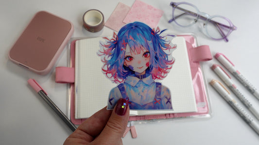 a hand holds a sticker of an anime girl with blue and red hair over a desk containing a pink hobonichi techo journal, mildliner pens in pink, pastel glasses, an inspic photo printer and a pink felt tip pen