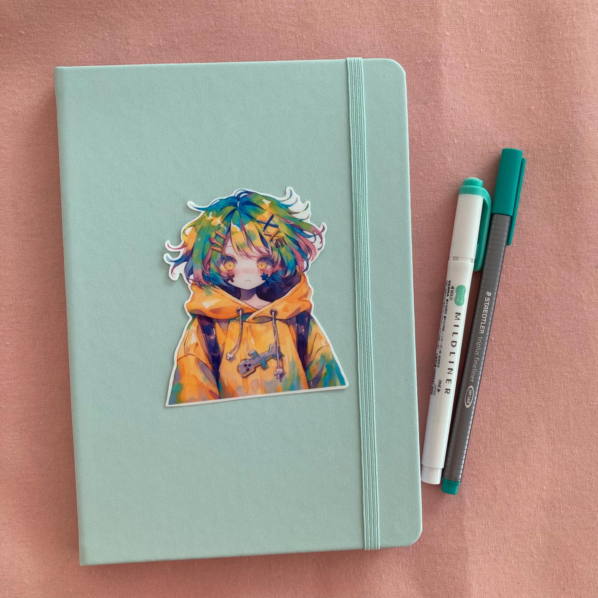A colorful anime sticker of a character with multicolored hair and a yellow hoodie is placed on a light green notebook with an elastic band. Next to the notebook are a teal Mildliner highlighter and a green Staedtler pen. The background is soft pink
