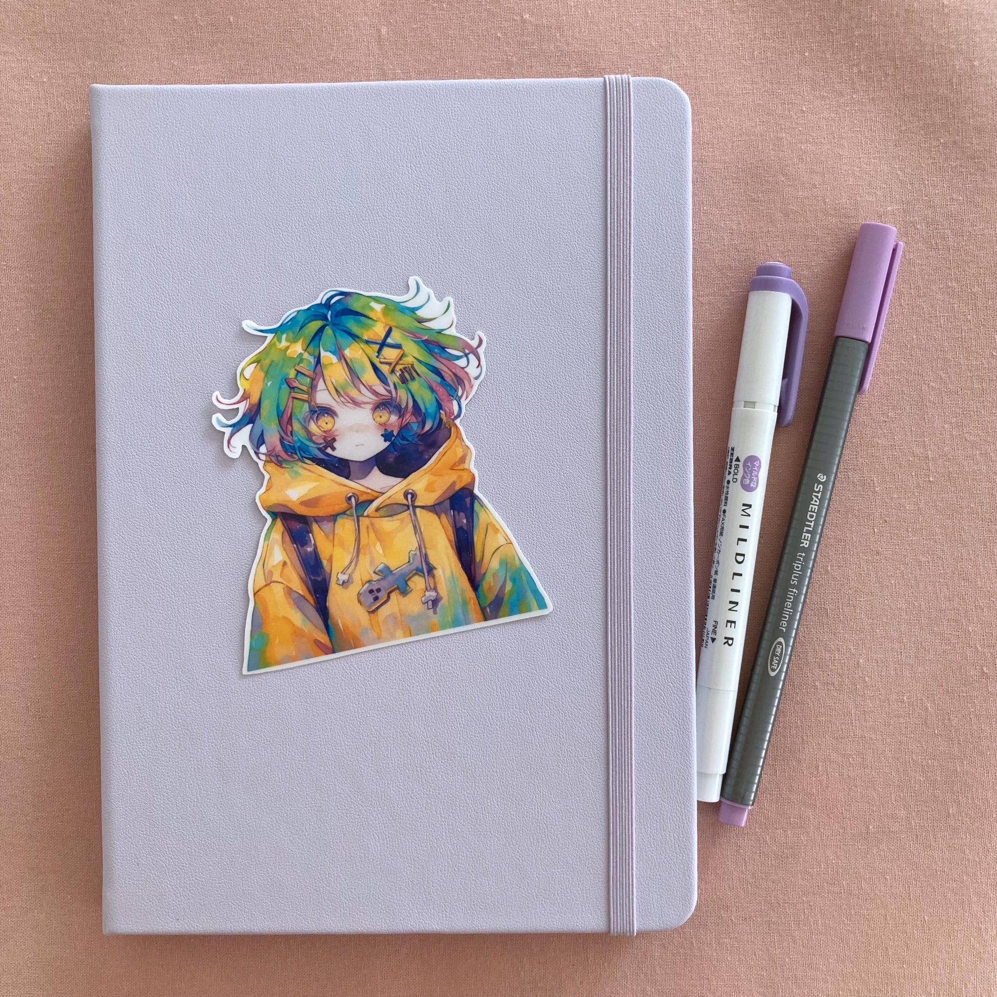 A colorful anime sticker of a character in a yellow hoodie and multicolored hair is placed on a light lavender notebook with an elastic band. Beside the notebook are a purple Mildliner highlighter and a gray Staedtler pen. The background is soft pink