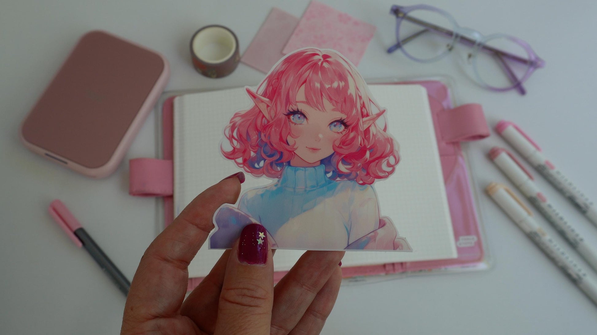 A hand holding a colorful anime sticker of a character with curly pink hair, bright eyes, and elf ears, wearing a turtleneck sweater. The background includes a pink planner, purple glasses, pink markers, washi tape, and a pink portable printer.