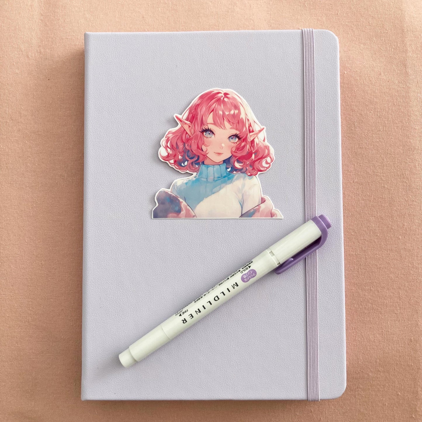A pastel lavender notebook with an anime sticker featuring a character with curly pink hair, elf ears, and bright eyes. A purple Mildliner highlighter rests diagonally on the notebook, all set against a soft pink fabric background.