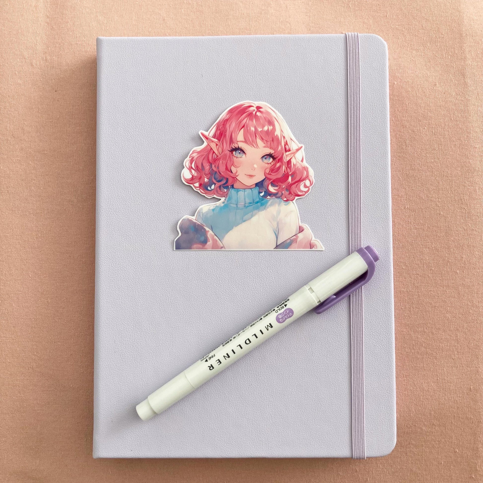 A pastel lavender notebook with an anime sticker featuring a character with curly pink hair, elf ears, and bright eyes. A purple Mildliner highlighter rests diagonally on the notebook, all set against a soft pink fabric background.