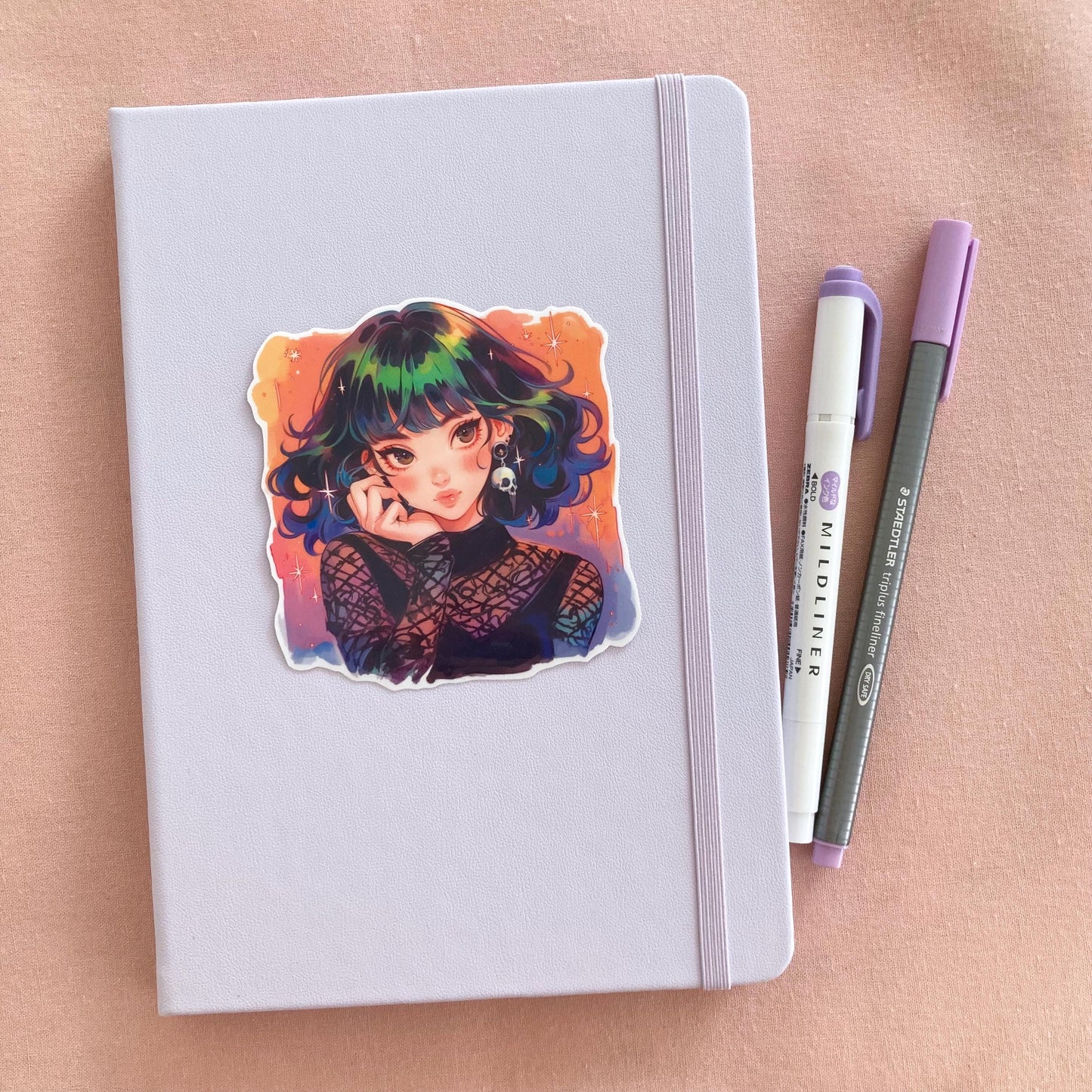 A pastel lavender notebook with a colorful anime sticker of a girl with rainbow hair, star accents, and a skull earring in a gothic mesh outfit. Next to the book are a purple Mildliner highlighter and a gray Staedtler pen on a pink fabric background