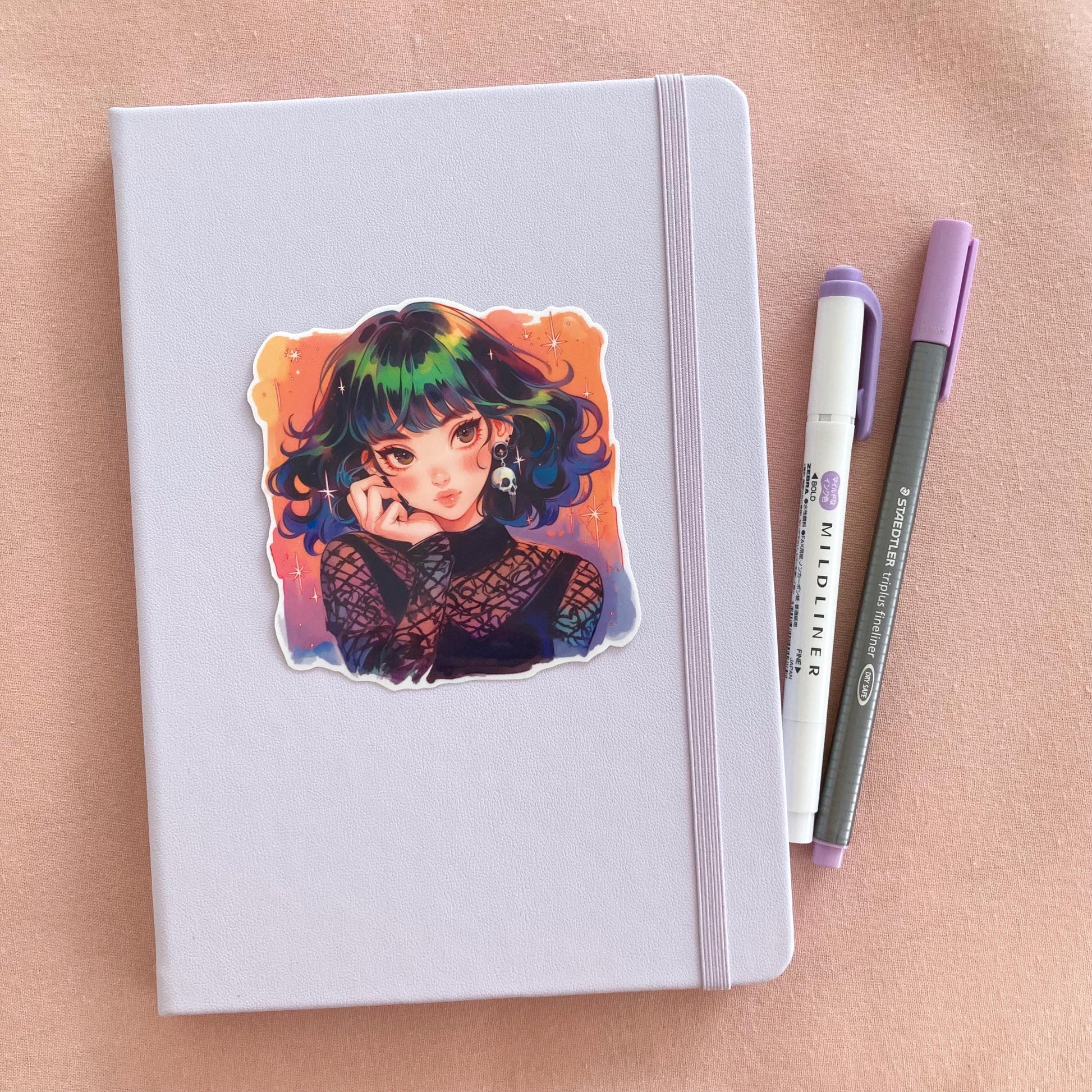 A pastel lavender notebook with a colorful anime sticker of a girl with rainbow hair, star accents, and a skull earring in a gothic mesh outfit. Next to the book are a purple Mildliner highlighter and a gray Staedtler pen on a pink fabric background