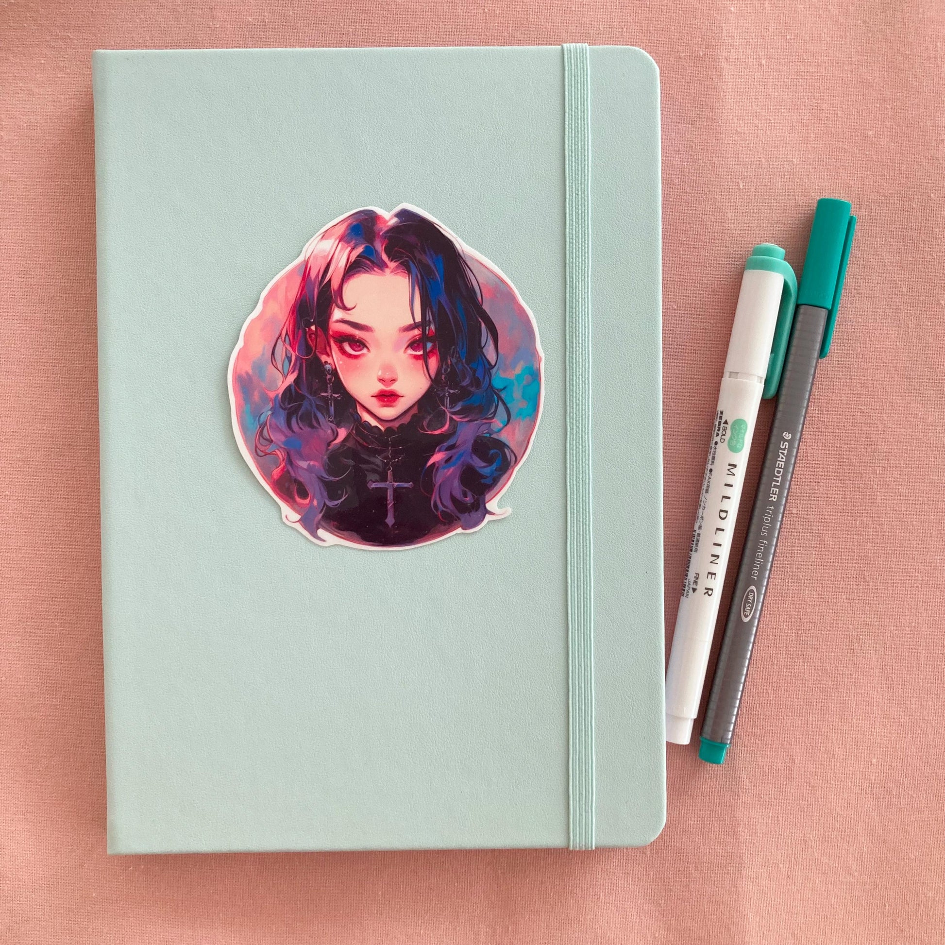 A pastel green notebook with a round sticker of an anime-style girl featuring long black hair with pink and blue highlights, wearing cross jewelry. To the right are a teal marker and pen, all placed on a light pink surface.