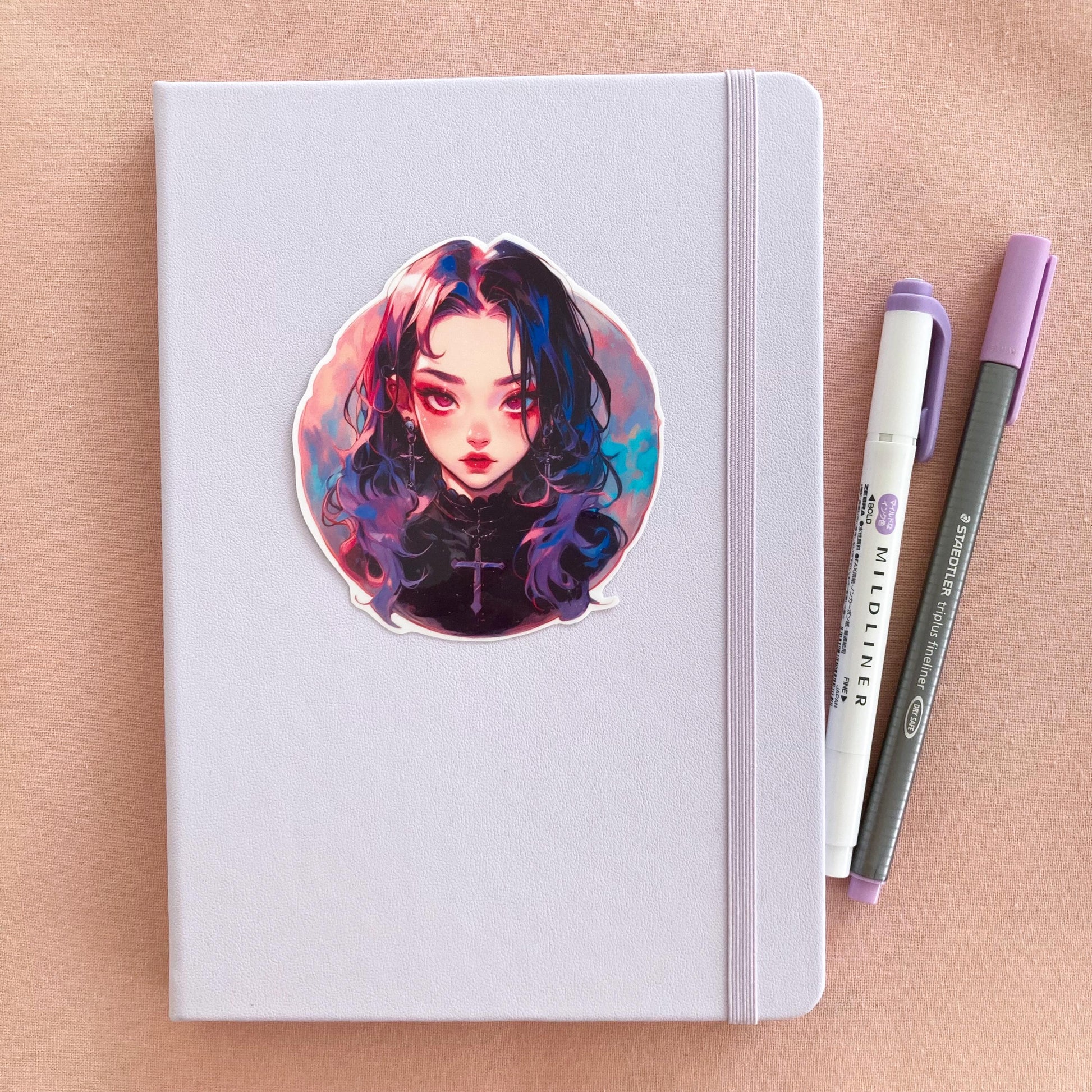 A pastel purple notebook with a round sticker of an anime-style girl with long black hair, pink and blue highlights, and cross jewelry. Beside it are a purple marker and pen, all placed on a light pink surface.