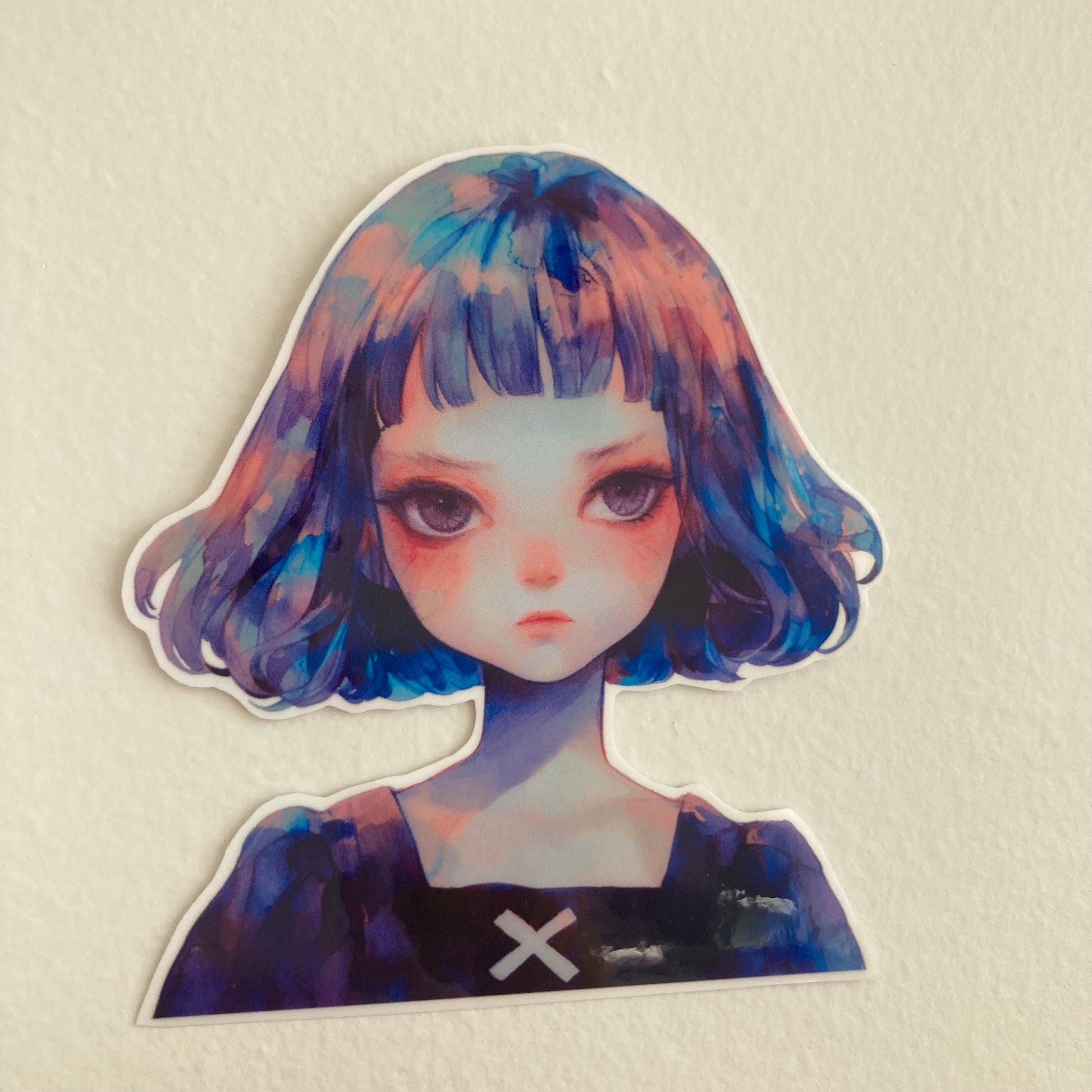 A sticker of a girl with short blue and purple hair, wearing a black top with a white &quot;X,&quot; placed against a plain light-colored surface.