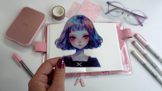 A hand holds a sticker of a girl with blue and purple hair, set against a pink notebook. Surrounding items include markers, washi tape, pink paper, a compact printer, and glasses. The person&#39;s nails are painted purple with a silver star accent.