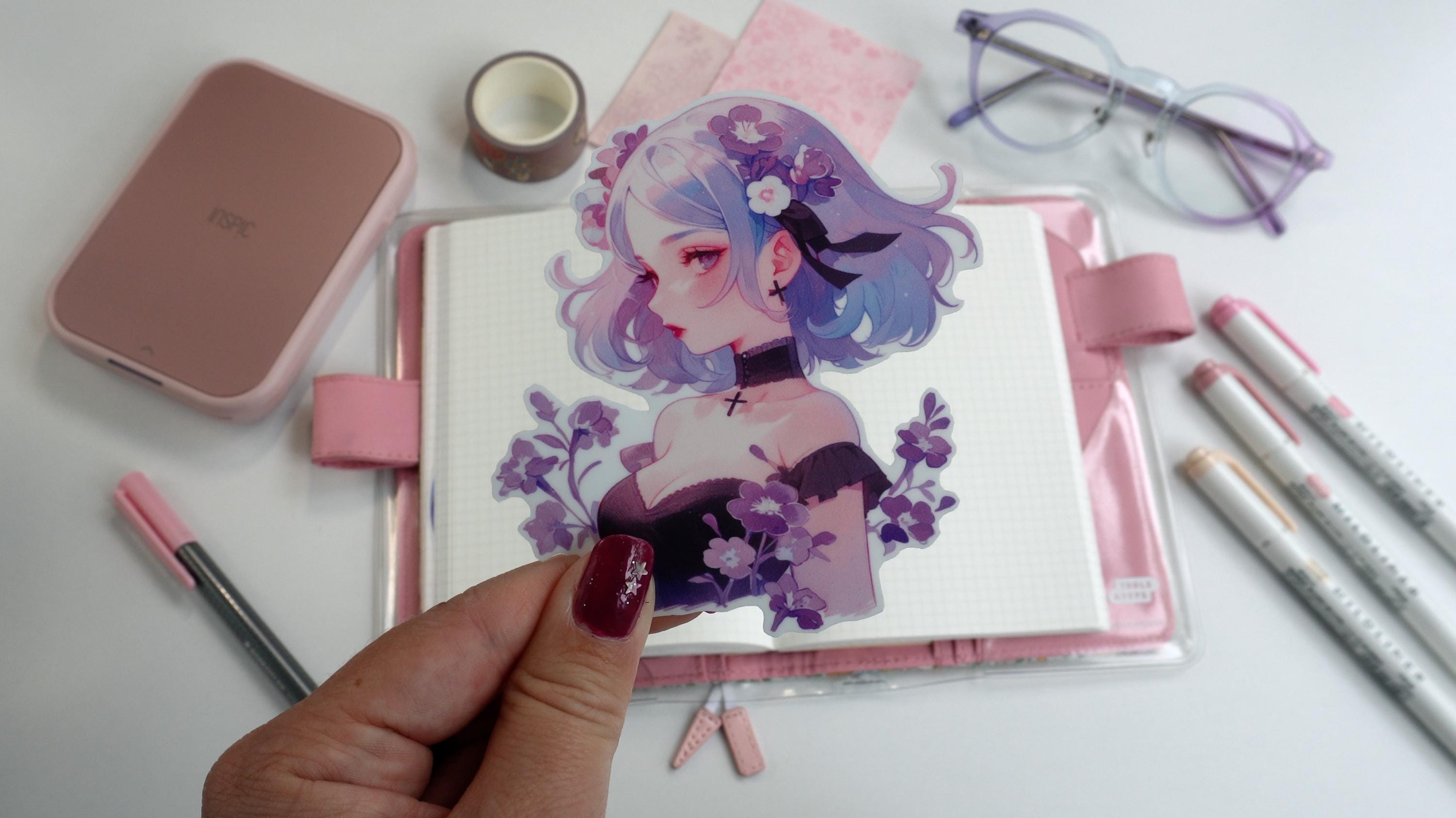 A hand holds a sticker of a girl with blue and purple hair with flowers, wearing a choker and black top. The background includes a pink book, markers, washi tape, paper, a printer, and glasses.