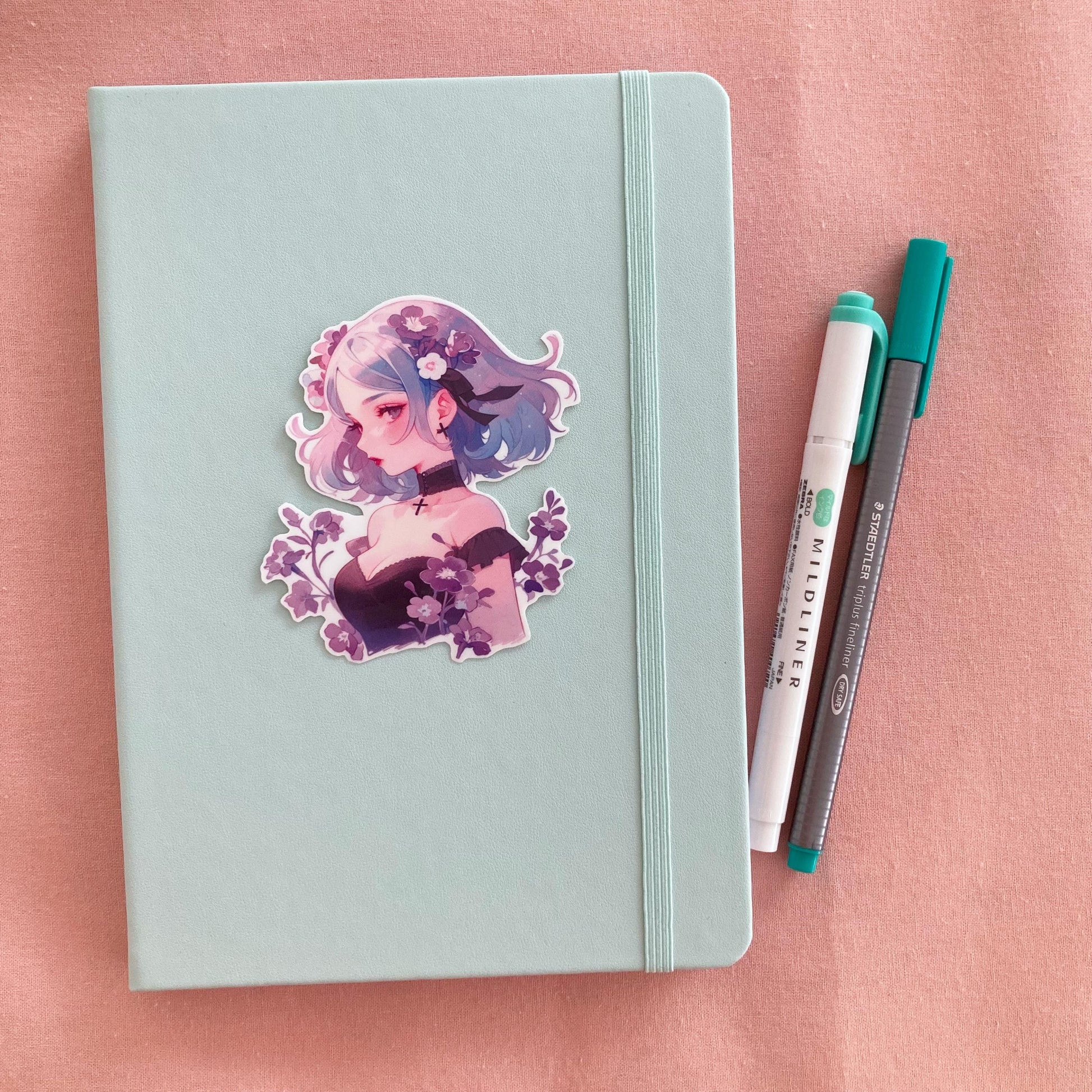 A sticker of a girl with blue and purple hair, surrounded by flowers, is placed on a light teal notebook. Beside the notebook are a green marker and a gray pen, all on a soft pink fabric background.