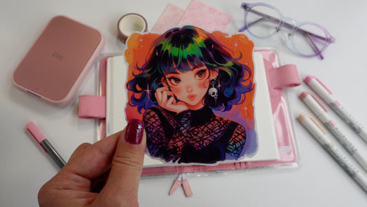 A hand holding a colorful anime sticker featuring a girl with rainbow hair, star accents, and a skull earring. She&#39;s wearing a gothic mesh outfit against a vibrant background. Behind are a pink planner, purple glasses, washi tape, and pink markers.