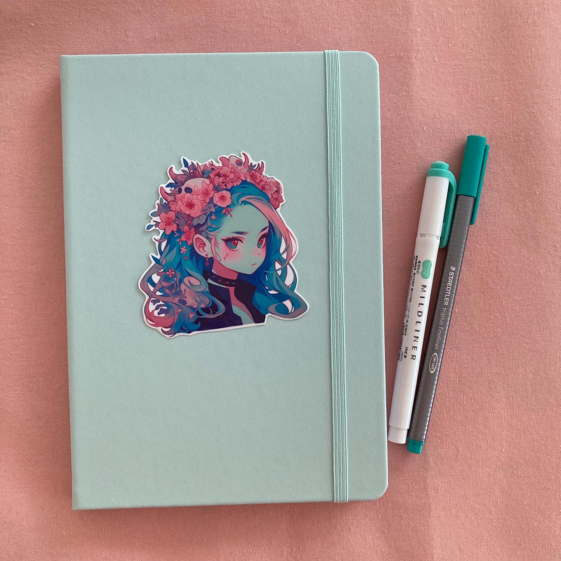 A sticker of a girl with blue hair and a floral crown is placed on a light teal notebook. Beside the notebook are a green marker and a gray pen, all on a soft pink fabric background.