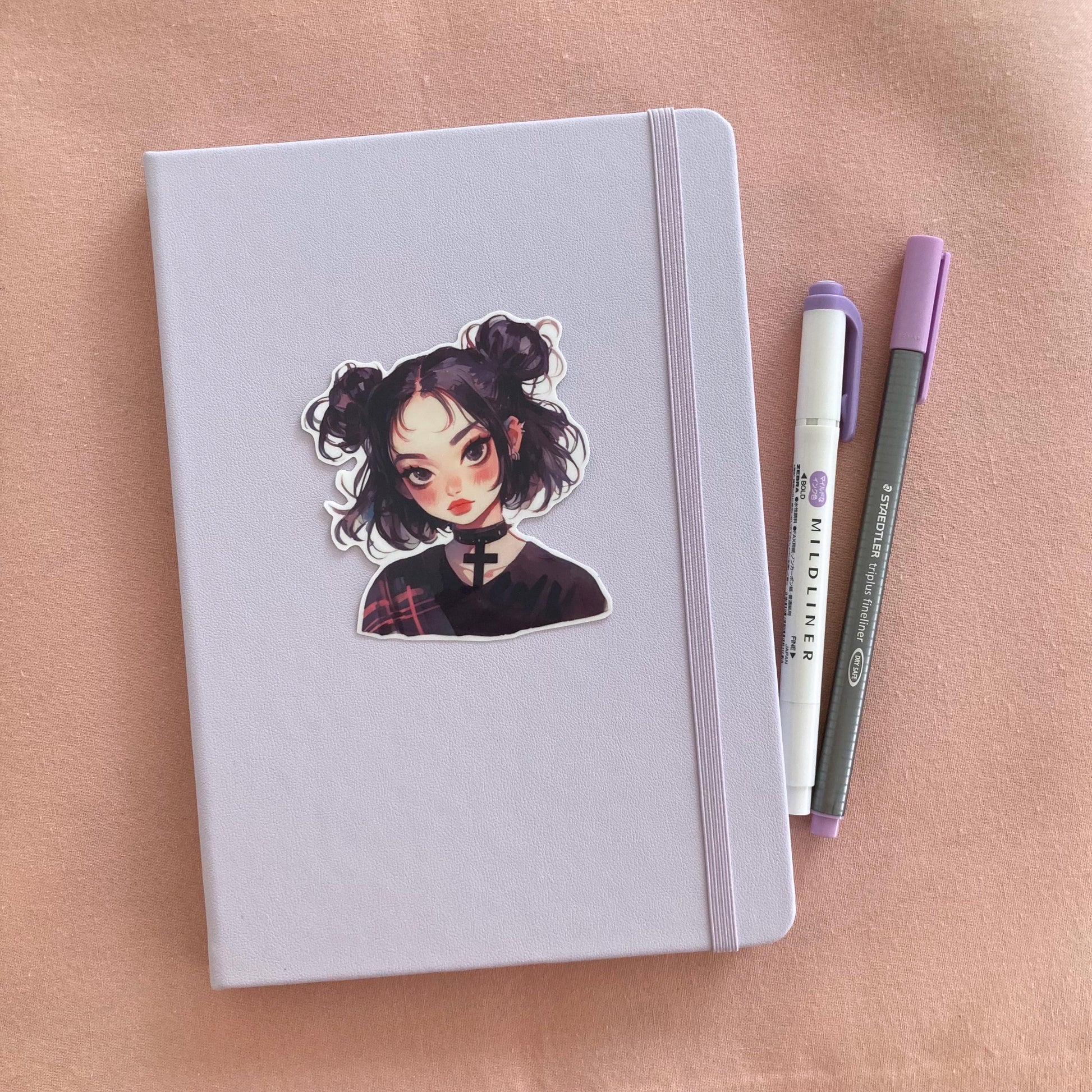 A sticker of a girl with black hair in buns and a choker is placed on a light lavender notebook. Beside it are a purple marker and a gray pen, all on a soft pink fabric background.