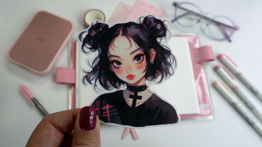A hand holds a sticker of a girl with short black hair in buns, wearing a choker and a dark shirt. The background features a pink book, markers, washi tape, pink paper, a printer, and glasses. The person&#39;s nails are dark pink with star accents.