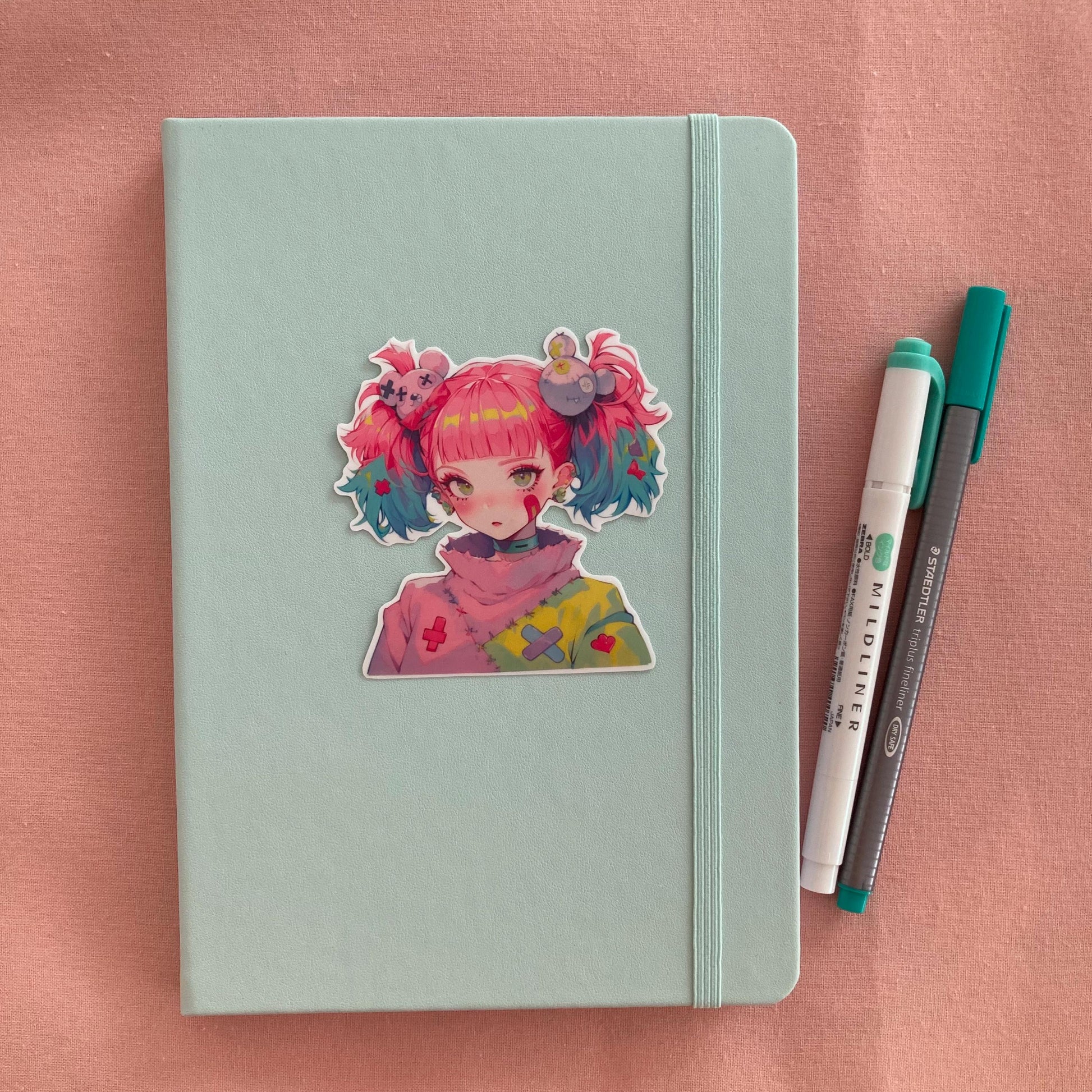 A pastel green notebook with an anime-style sticker of a girl with pink and blue pigtails on its cover, next to a green marker and gray pen, all set on a pink fabric background.