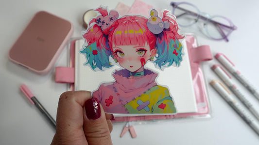 A hand holding a colorful anime-style sticker of a girl with pink and blue pigtails, wearing a pastel sweater. Background includes pink stationery, an open notebook, pastel markers, and glasses on a white surface.