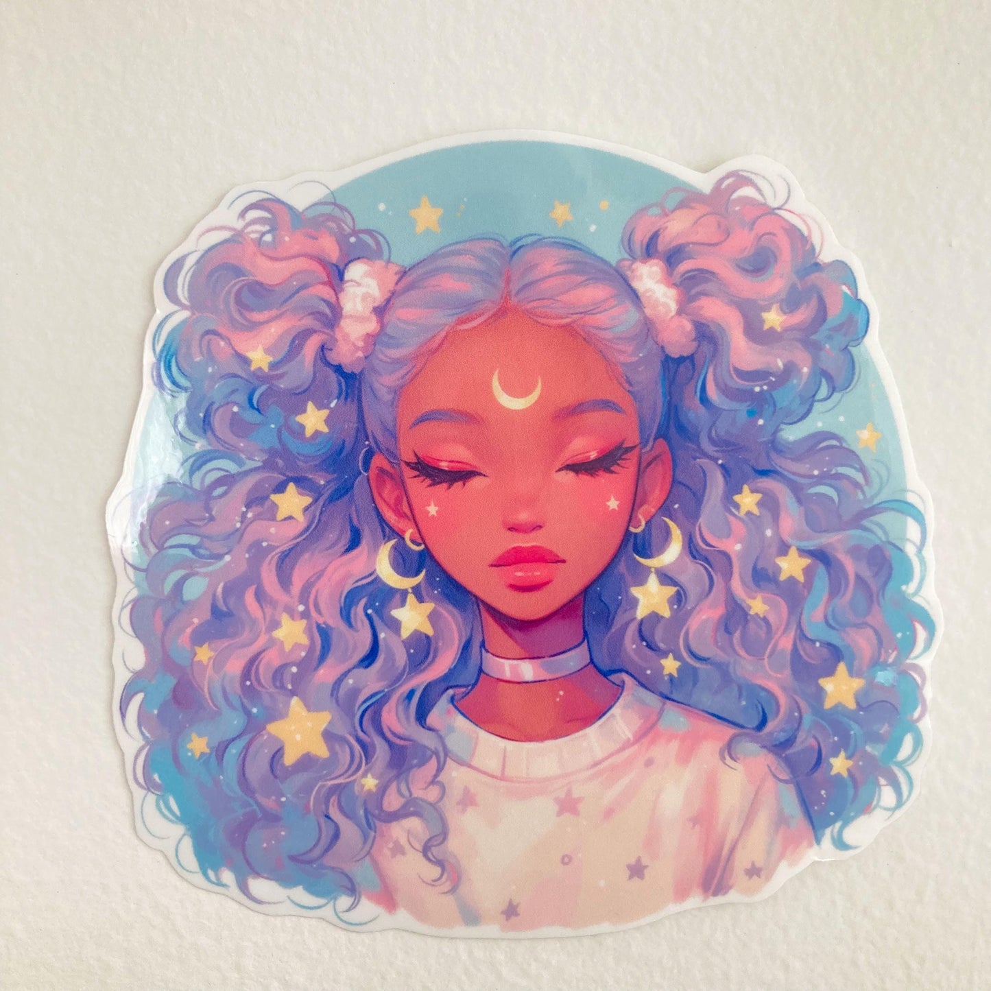 A colorful sticker of an anime-style girl with closed eyes, lavender and blue curly hair, adorned with stars, and a crescent moon on her forehead, placed on a plain white surface.