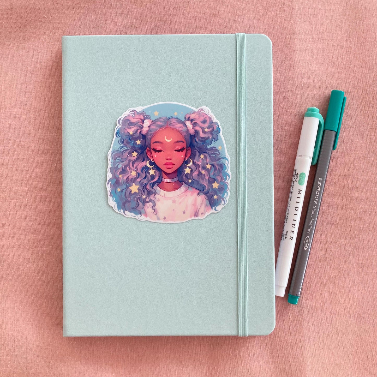 A pastel green notebook with an anime-style sticker of a girl with lavender and blue curly hair on its cover, placed next to a green marker and gray pen on a pink fabric background.