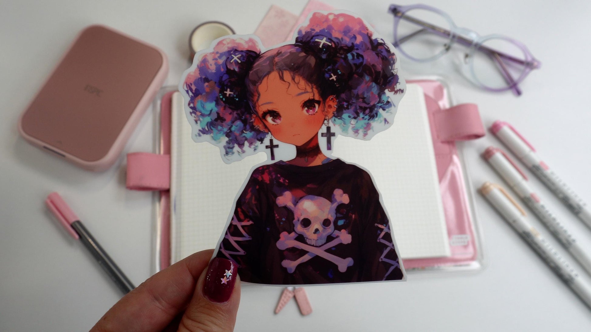 A hand holding a sticker of an anime-style character with purple and blue curly hair in pigtails, wearing cross earrings and a skull-themed shirt. The background includes pink stationery, an open notebook, and pastel markers on a white surface.