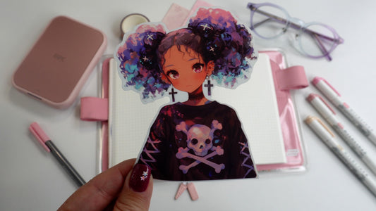 A hand holding a sticker of an anime-style character with purple and blue curly hair in pigtails, wearing cross earrings and a skull-themed shirt. The background includes pink stationery, an open notebook, and pastel markers on a white surface.