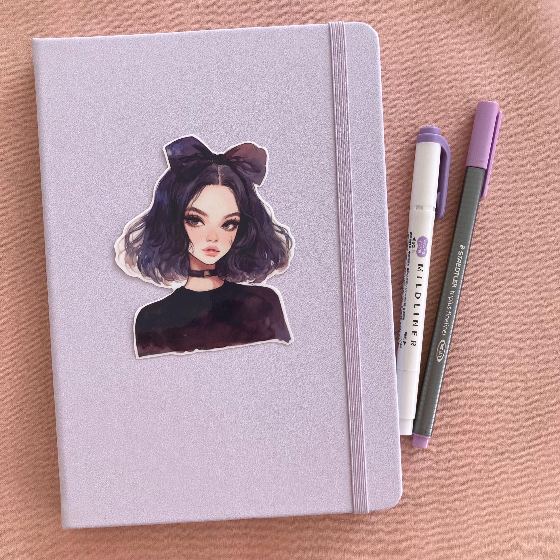 A pastel lavender notebook with a sticker of an anime-style character with dark purple hair, a large bow, and a black choker, next to a purple marker and gray pen on a pink fabric background.