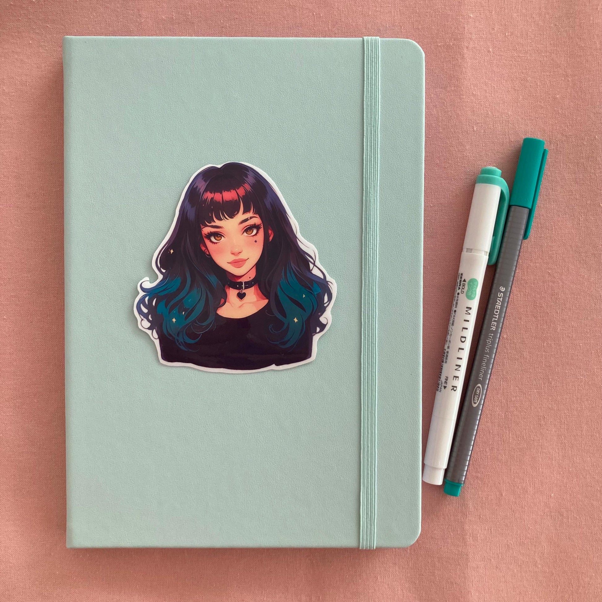 A pastel green notebook with a sticker of an anime-style character with black-to-teal hair and a heart choker, next to a green marker and gray pen, set on a pink fabric background.