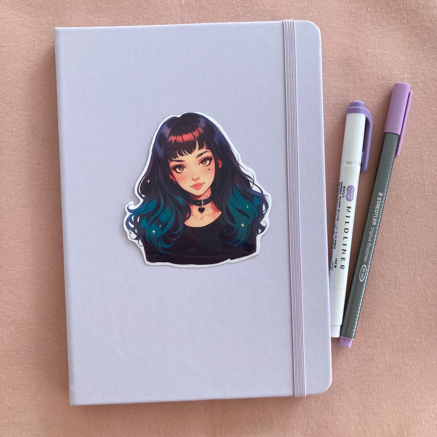 A pastel lavender notebook with a sticker of an anime-style character with black-to-teal hair and a heart choker, next to a purple marker and gray pen, set on a pink fabric background.