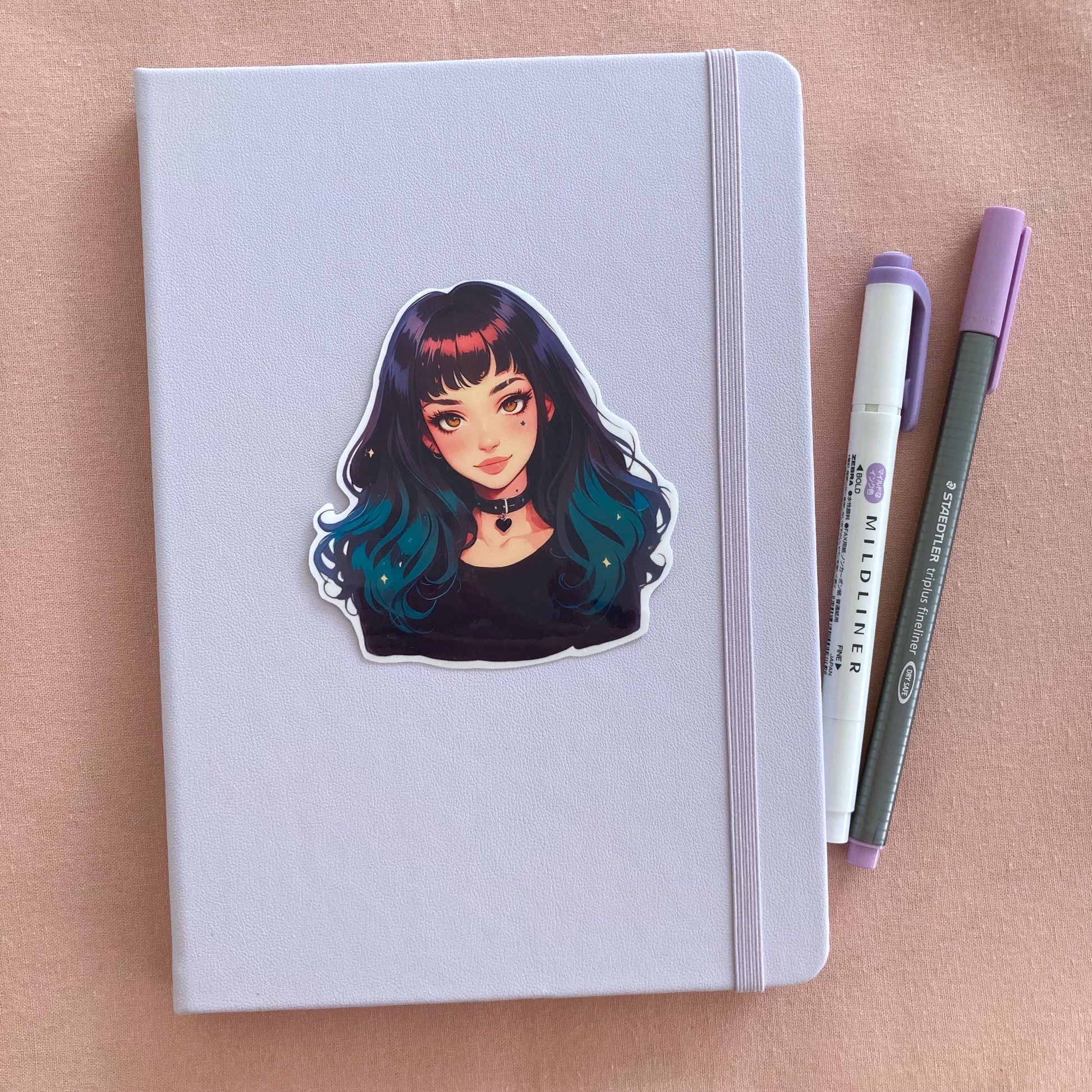 A pastel lavender notebook with a sticker of an anime-style character with black-to-teal hair and a heart choker, next to a purple marker and gray pen, set on a pink fabric background.