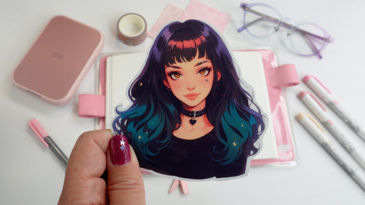 A hand holding a sticker of an anime-style character with long black hair fading to teal, a heart choker, and bright eyes. Background includes pink stationery, an open notebook, pastel markers, and glasses on a white surface.