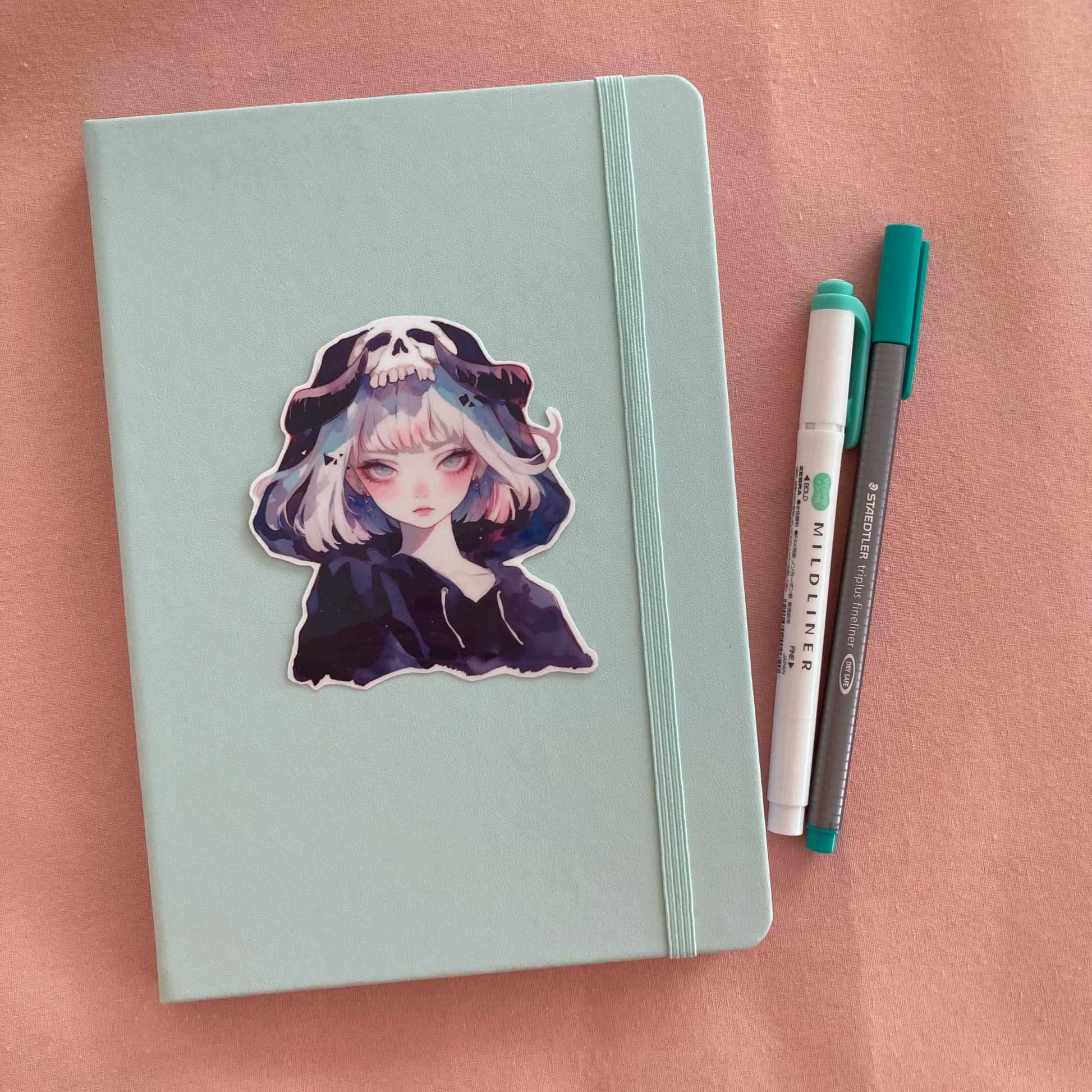 A pastel green notebook with a sticker of an anime-style character wearing a hood with a skull design, next to a green marker and gray pen, set on a pink fabric background.