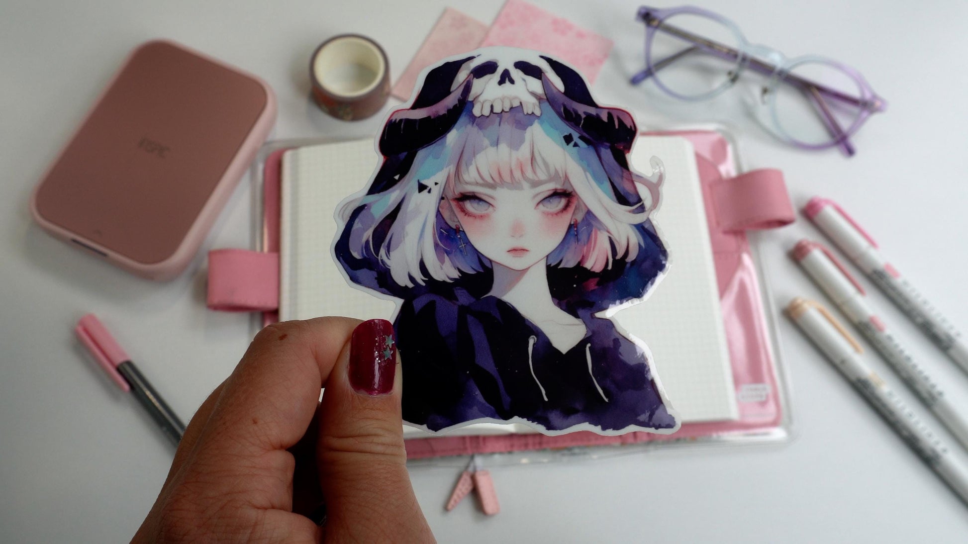 A hand holding a sticker of an anime-style character with short silver hair, wearing a hood with a skull design. Background includes pink stationery, an open notebook, pastel markers, and glasses on a white surface.