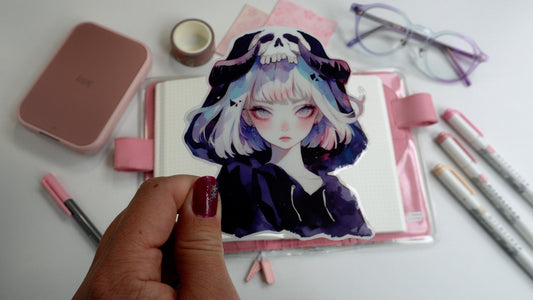 A hand holding a sticker of an anime-style character with short silver hair, wearing a hood with a skull design. Background includes pink stationery, an open notebook, pastel markers, and glasses on a white surface.