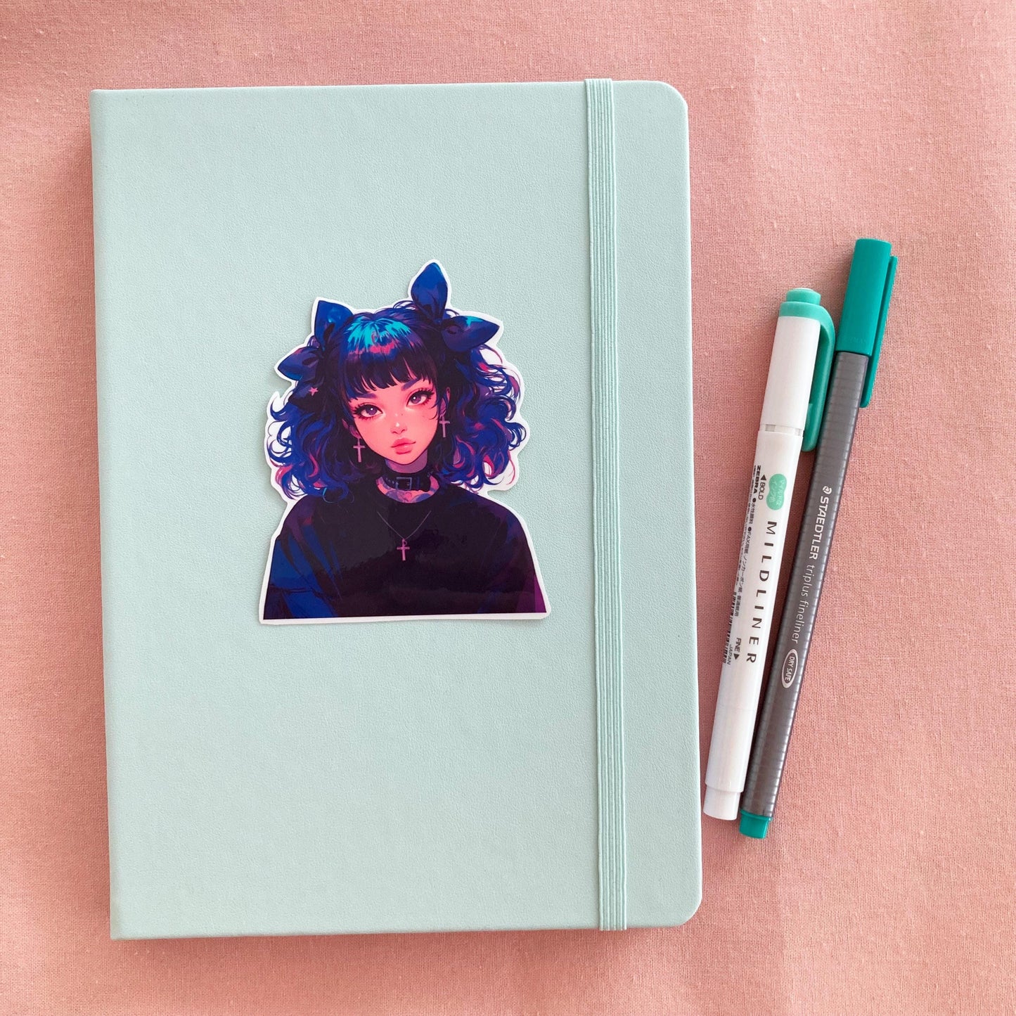 A pastel green notebook with a sticker of an anime-style character with blue and pink curly hair, large bows, and cross earrings, next to a green marker and gray pen on a pink fabric background.