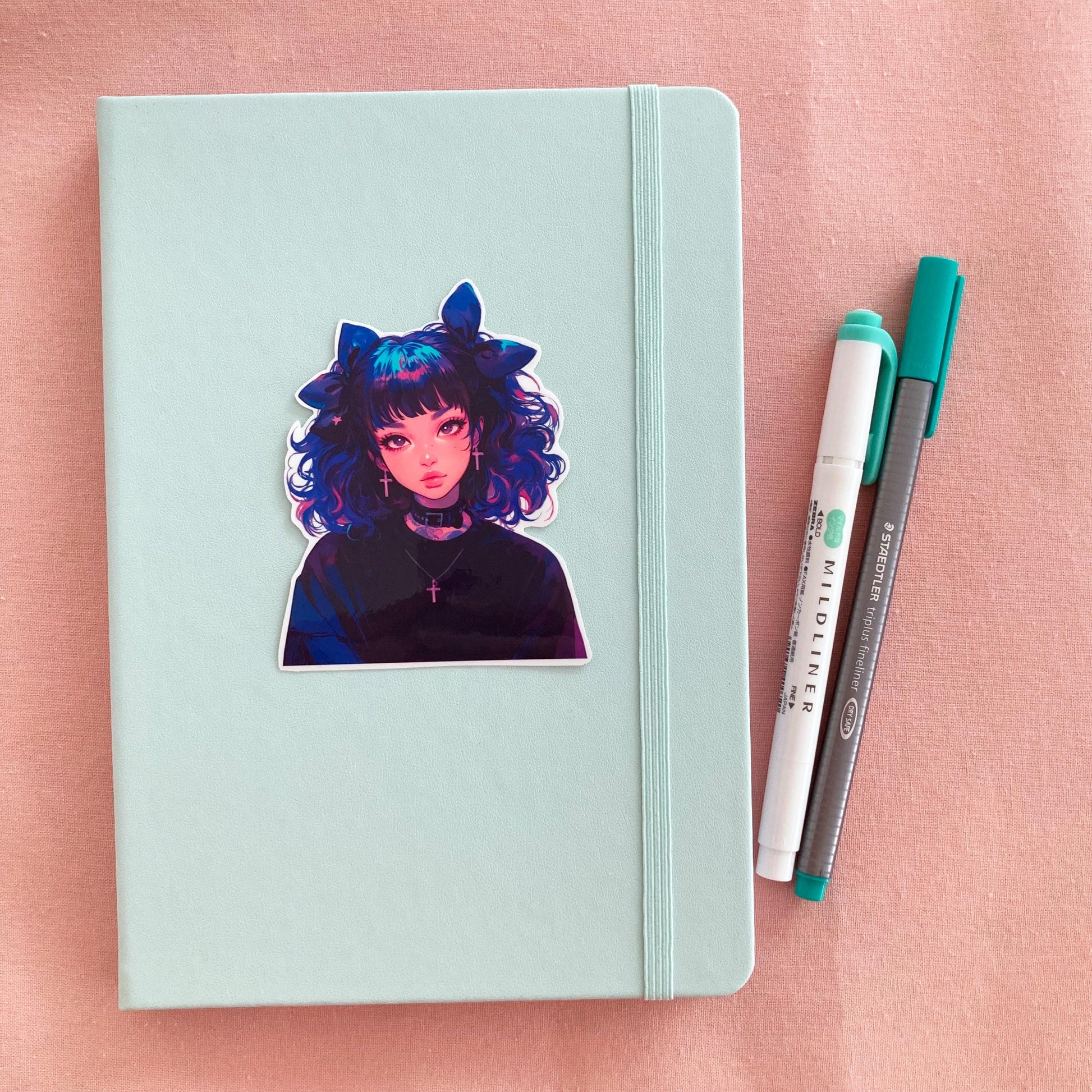 A pastel green notebook with a sticker of an anime-style character with blue and pink curly hair, large bows, and cross earrings, next to a green marker and gray pen on a pink fabric background.