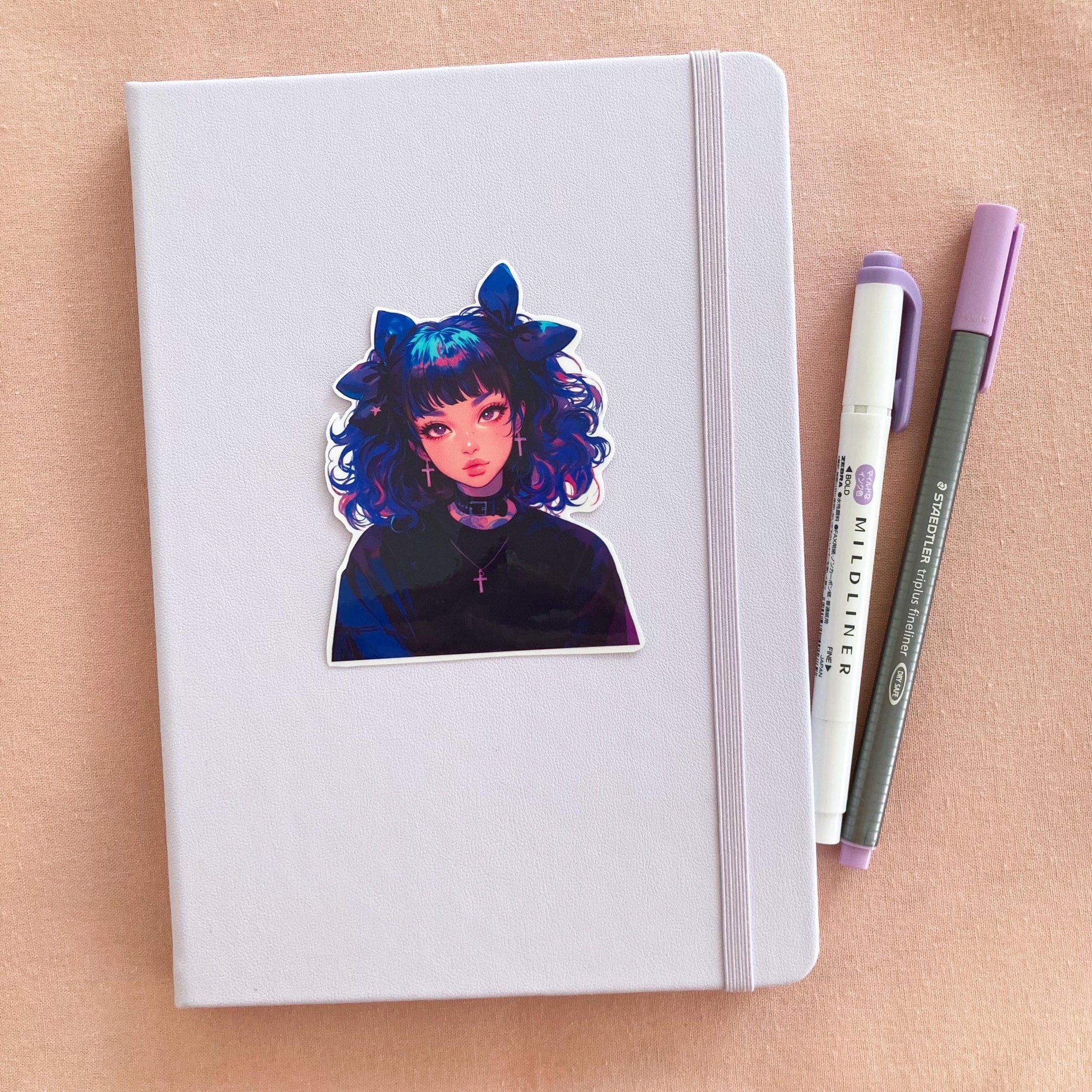 A pastel lavender notebook with a sticker of an anime-style character with blue and pink curly hair, large bows, and cross earrings, next to a purple marker and gray pen on a pink fabric background.