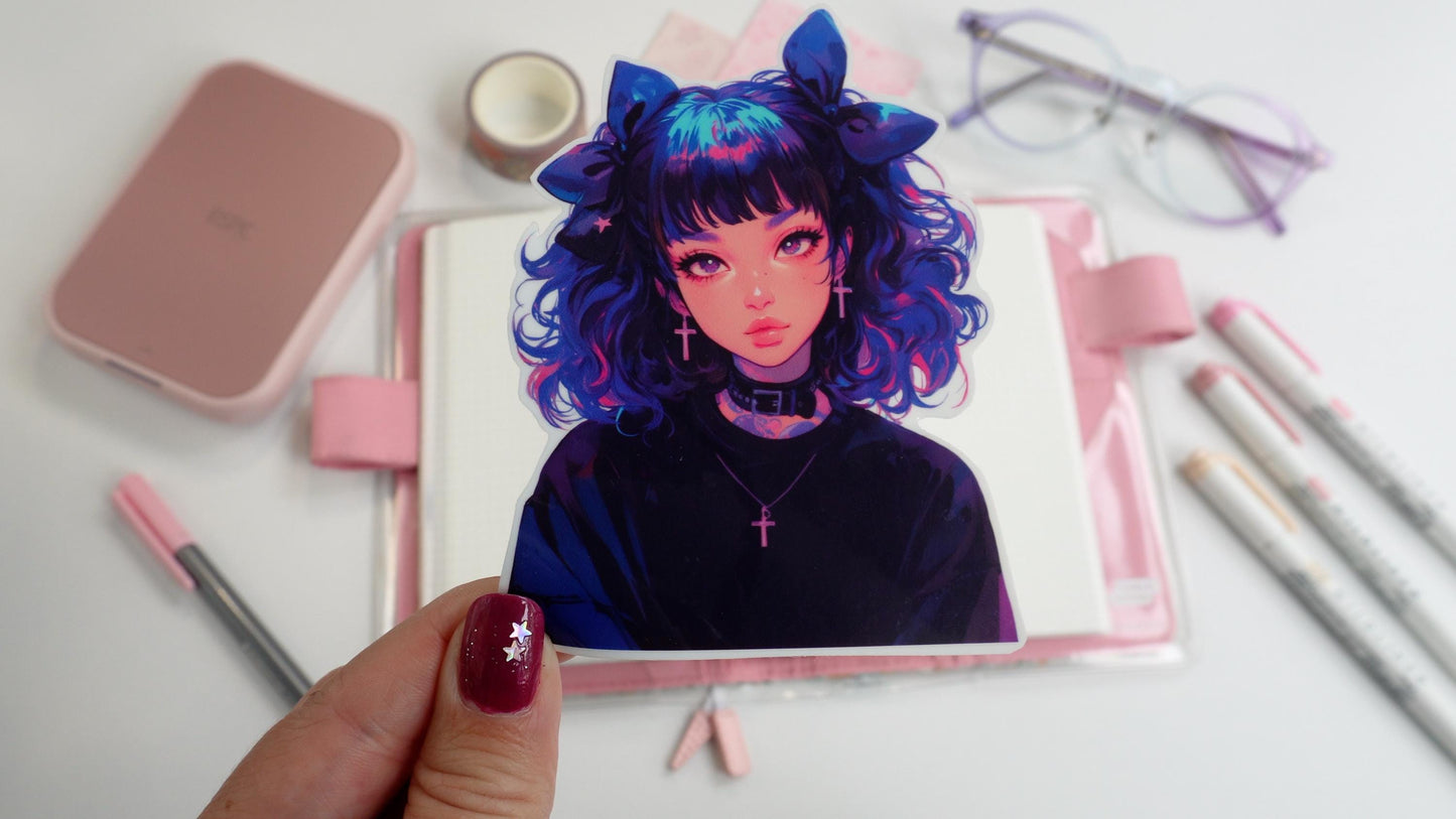 A hand holding a sticker of an anime-style character with curly blue and pink hair, large bows, and cross earrings, wearing a black choker. Background includes pink stationery, an open notebook, pastel markers, and glasses on a white surface.
