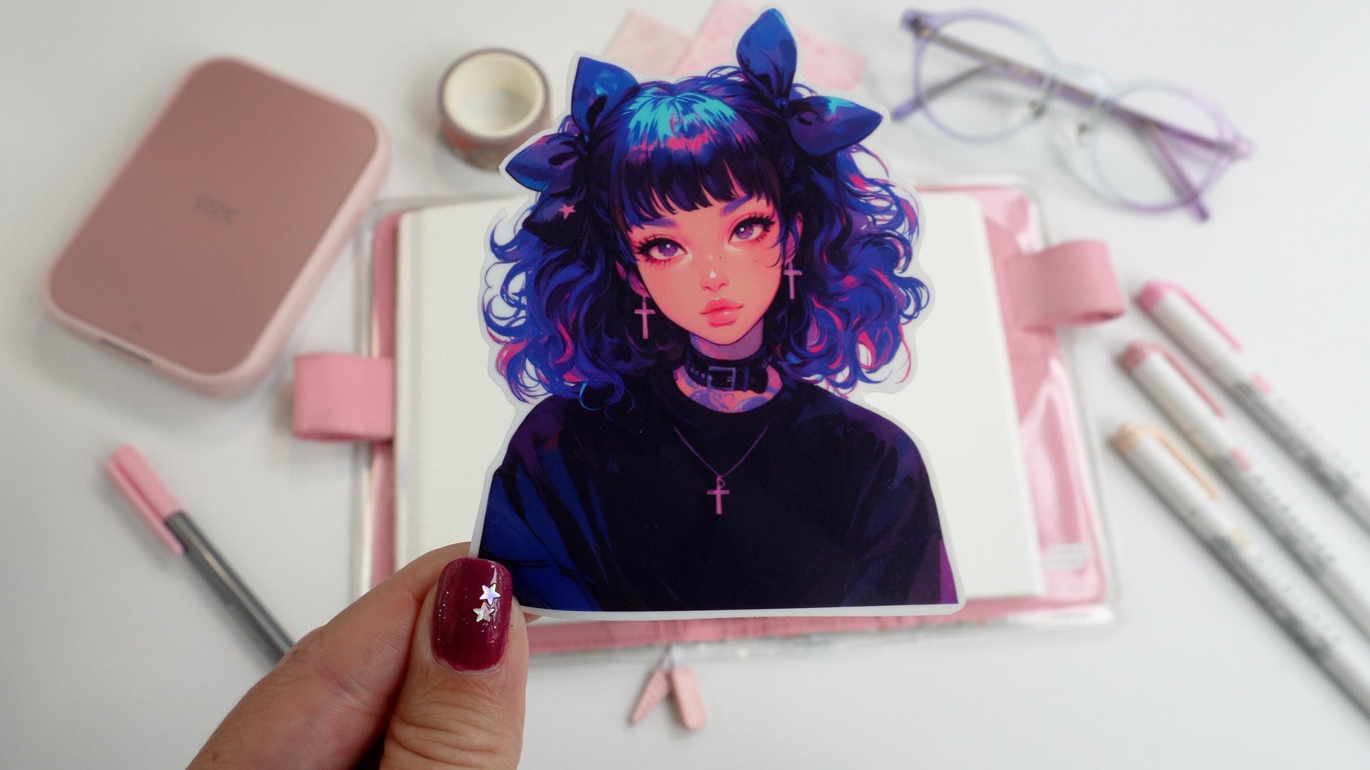 A hand holding a sticker of an anime-style character with curly blue and pink hair, large bows, and cross earrings, wearing a black choker. Background includes pink stationery, an open notebook, pastel markers, and glasses on a white surface.