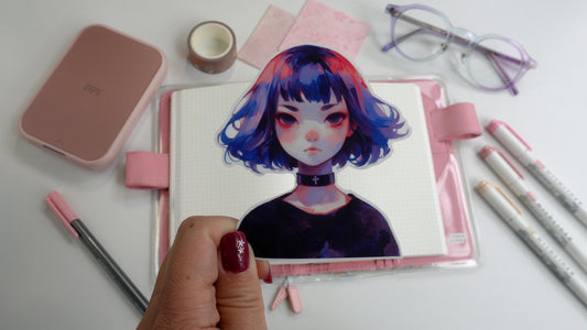 A hand holding a sticker of an anime-style character with a short blue and pink bob haircut, wearing a black choker with a cross. Background shows pink stationery items, a planner, and markers, with the hand&#39;s nail painted purple with star accents.