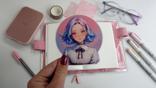 A sticker held by hand of an anime-style character with blue and pink hair, wearing a white blouse with a black ribbon. In the background are stationery items: pastel-colored pens, glasses, washi tape, and a pink notebook with a grid pattern.