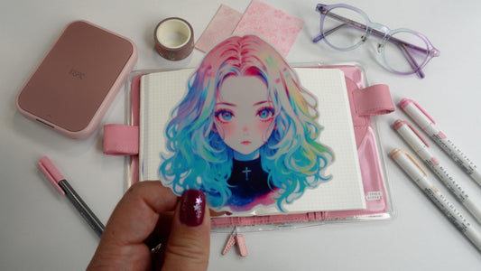 A hand holding a colorful anime sticker of a girl with rainbow hair over a notebook. Surrounding items include pink pens, glasses, a pink device, decorative tape, and pink papers on a white surface. Nails are painted purple with star designs.