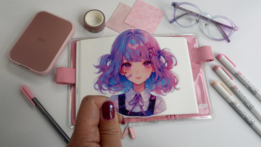 A hand with maroon nail polish and a silver star holds a sticker of a girl with blue and pink hair. The background includes a notebook, pink case, markers, glasses, washi tape, and pink paper on a white surface.