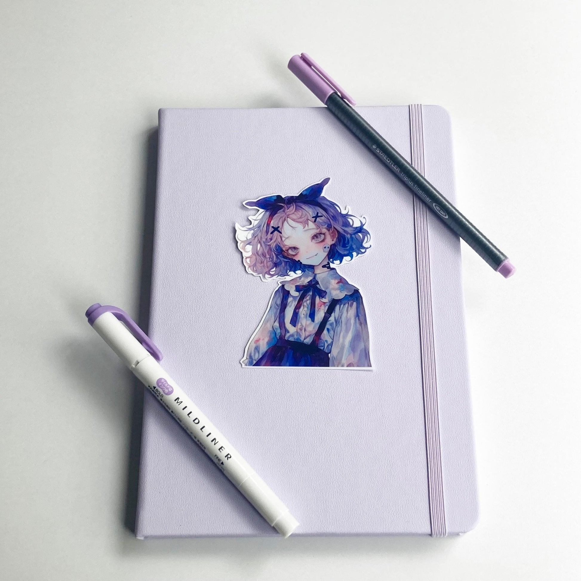 A sticker of an anime-style character is placed on a closed lavender notebook with an elastic band. Two pens, one gray with a purple cap and one white &quot;Mildliner&quot; marker with a purple cap, are laid diagonally across the notebook.