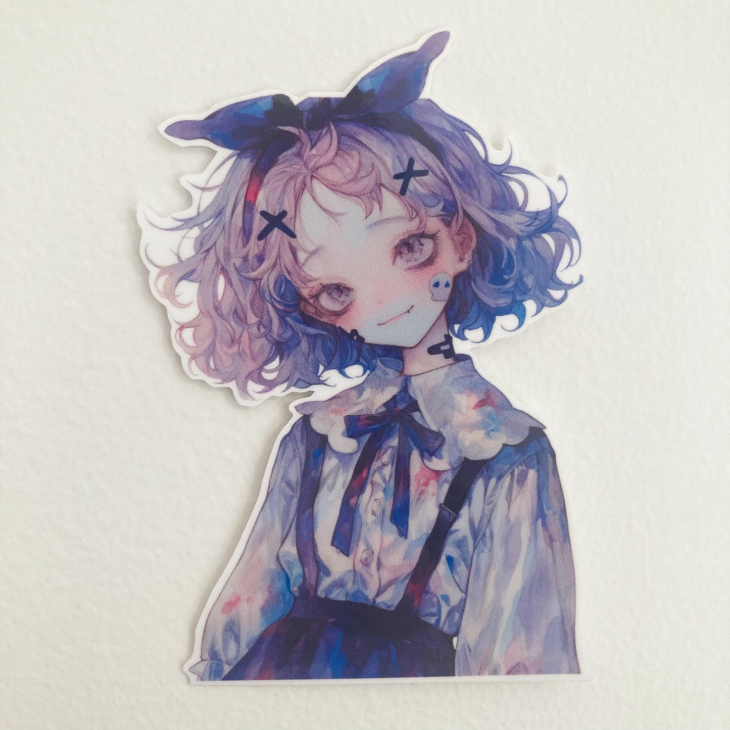 A sticker of an anime-style character with pastel-colored hair, a blue bow, and a detailed outfit is placed on a textured white surface. The character has a slight smile, expressive eyes, and a soft color palette in blue and purple hues.
