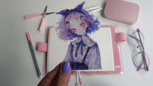 A hand with purple polish holds a sticker depicting an anime character with pastel hair and a bow. The background includes a pink planner, pink and gray pens, pink scissors, purple glasses, and a pink case, all arranged on a white surface