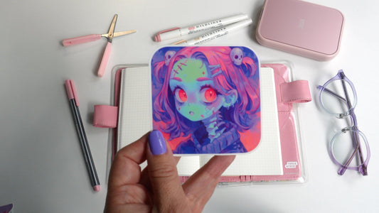 A hand with purple nails holds a square sticker of a colorful anime character with pink hair, red eyes, and a skull accessory. The background features a pink notebook, pens, pink scissors, a pink case, and purple-rimmed glasses on a white surface.