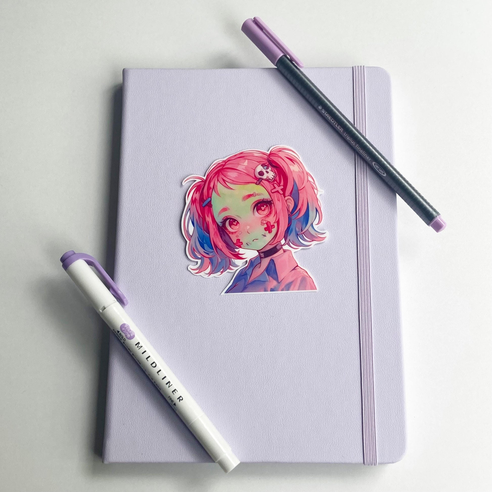 A sticker of an anime character with pink and blue hair is placed on a closed light purple notebook. A purple marker and a pen with a purple cap lie diagonally across the notebook.